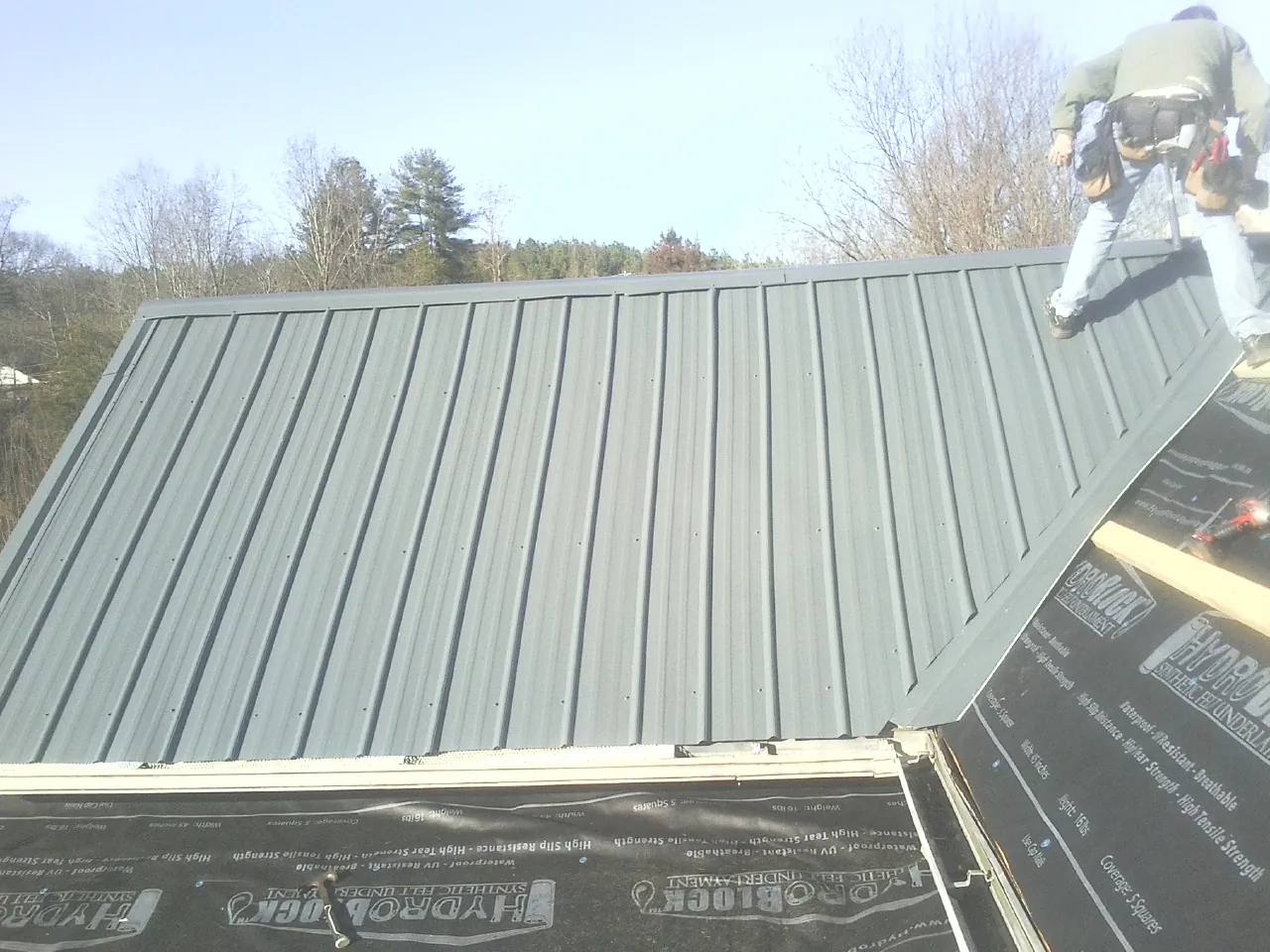 Crew installing dark green standing seam panels over synthetic underlayment in Pineola