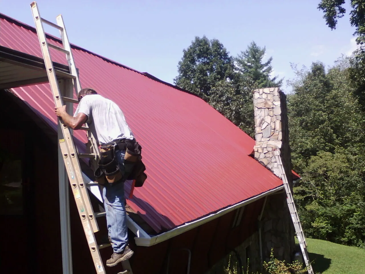 Roofer on a ladder beside a red metal roof and stone chimney — Soffit & Fascia Repair by Roofing Brothers in Pineola